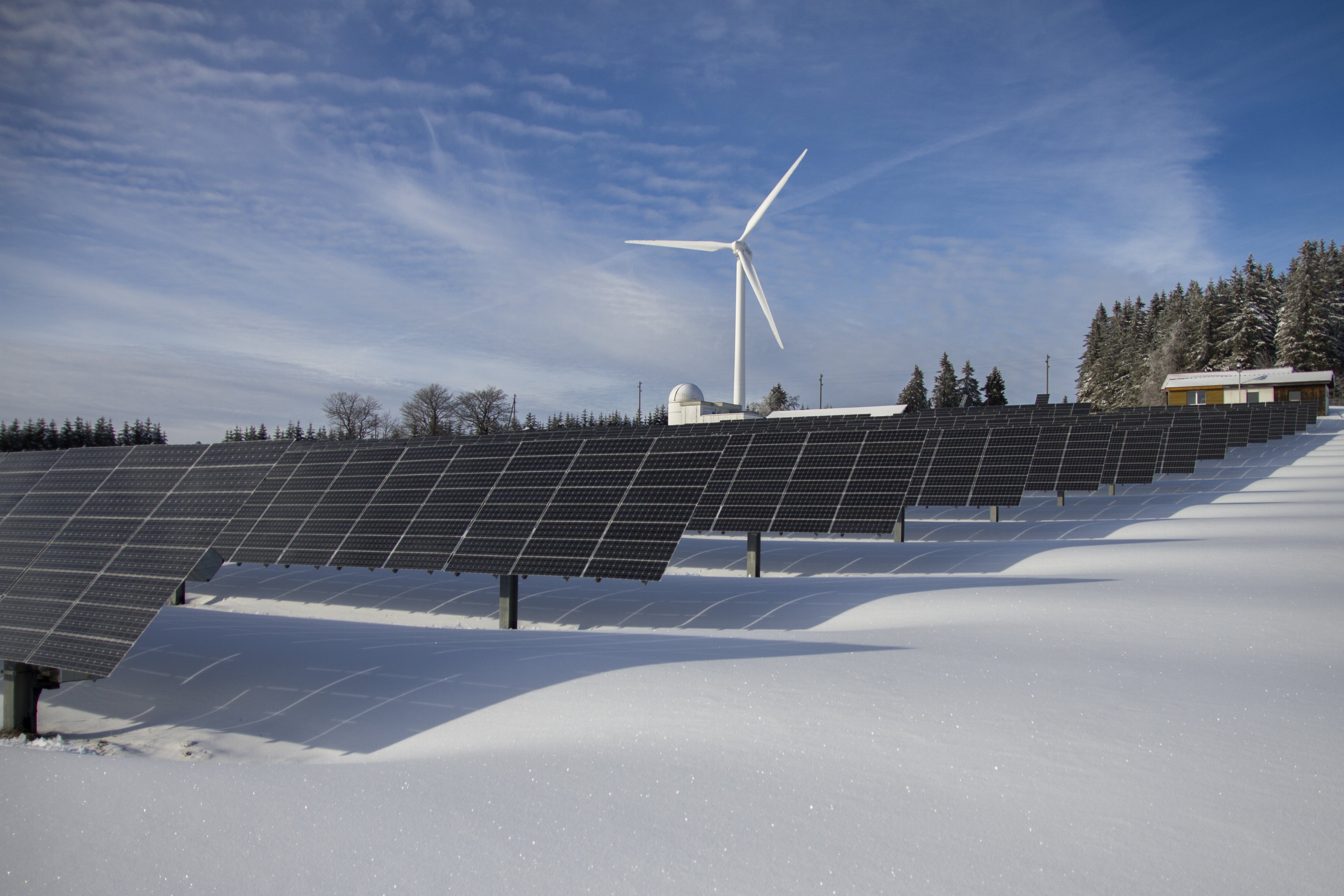 Solar panels on snow with windmill under clear day sky.